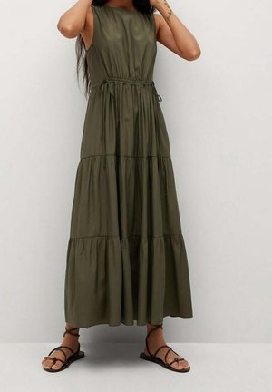 Femme portant une robe longue à volants sans manches vert olive avec taille à cordon et sandales plates noires à brides.