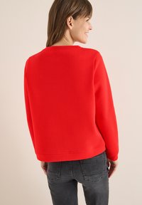 Felpa rossa con scollo rotondo e vestibilità morbida, realizzata in tessuto morbido. Accompagnata da jeans grigio scuro con dettagli sulle tasche.