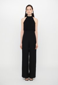 Svart halter jumpsuit med vide ben, glatt tekstur, rynket bodice og en smal midje. Enkel design kombinert med svarte sandaler.
