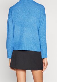 Blauer Strickpullover mit geripptem Ausschnitt und Saum. Kurze, leicht oversized Ärmel und eine lockere Passform. Tragen über einem schwarzen Rock.