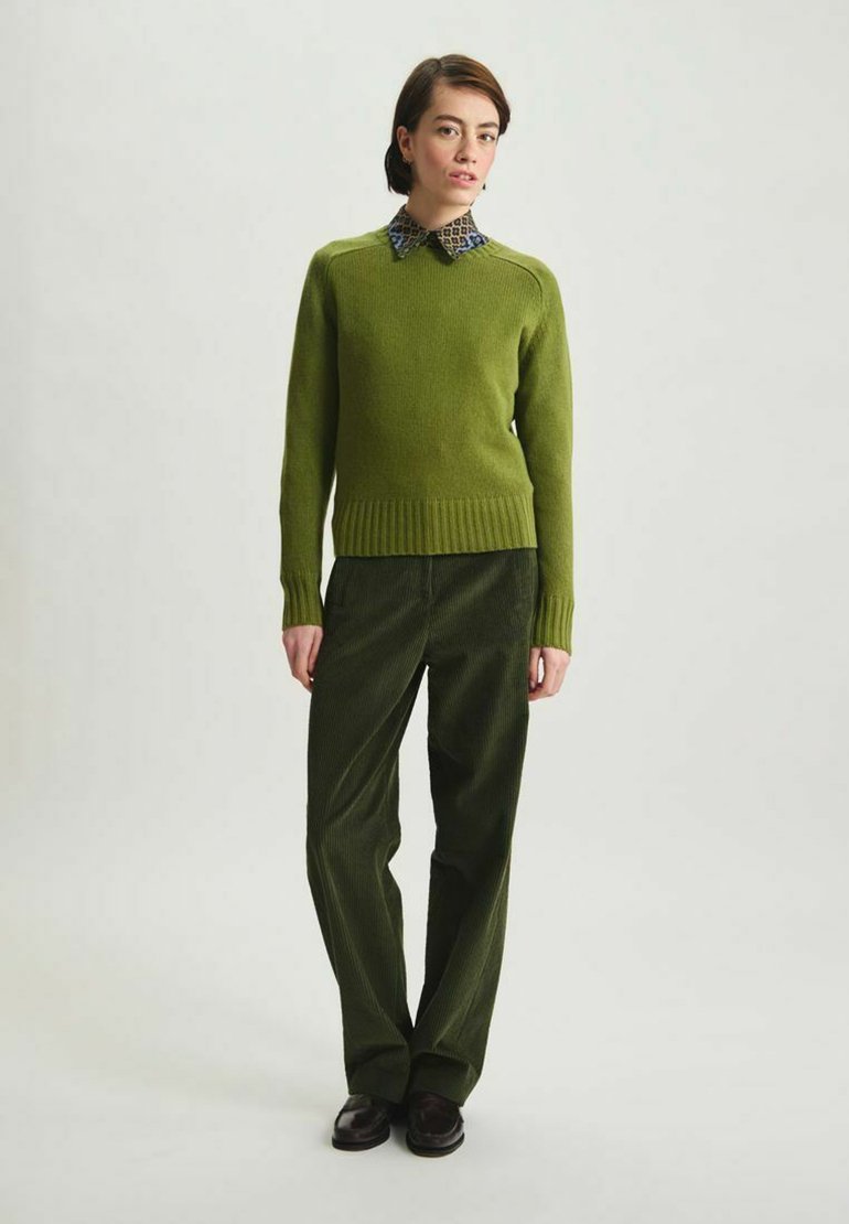 Un pull en maille vert avec un ourlet côtelé, associé à un pantalon en velours côtelé vert foncé. La tenue comporte une chemise à col à motifs en dessous.