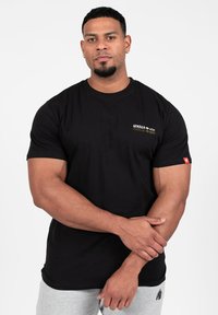 Juodas trumpomis rankovėmis medvilnės marškinėliai su auksiniu logotipu krūtinėje ir raudonu akcento ženkleliu ant rankovės. Paprastas dizainas.