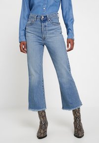 Light blue high-waisted jeans with a flared leg, featuring a raw hem and classic five-pocket design, paired with patterned ankle boots.