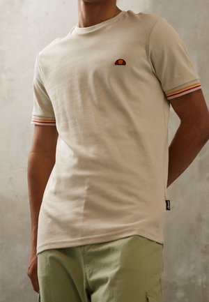 Mand iført beige t-shirt med korte ærmer med orange og gule stribede ærmekanter og lille logo på venstre bryst, kombineret med lysegrønne bukser.