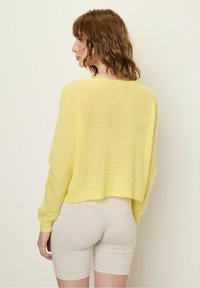 Gelbes, kurzes Chenille-Pullover mit langen Ärmeln. Leichte, weiche Textur. Über beigen Shorts getragen, die eine lockere Passform und ein offenes Rückendesign zeigen.