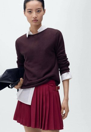 Pull en tricot bordeaux superposé à une chemise blanche à col, associé à une jupe plissée rouge avec un détail de boucle sur le côté. Sac à main noir porté dans la main.