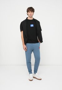 Juoda marškinėliai su mėlynu logotipu, derinami su mėlynomis laisvalaikio kelnėmis, kurios turi raudoną akcentą. Modelis dėvi baltus sportbačius su rudomis padais.