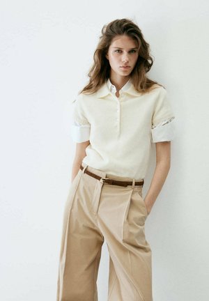 Joven mujer con cabello largo que lleva una camisa blanca de manga corta y pantalones beige de cintura alta con un cinturón marrón apoyada contra una pared blanca.