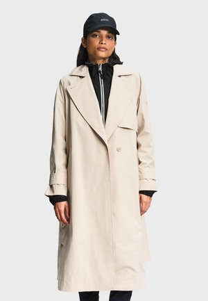 Žena u dugom bež baloneru preko crne jakne na patentni zatvarač i crnoj kapi s natpisom "AIGLE", stoji na jednobojnoj pozadini.