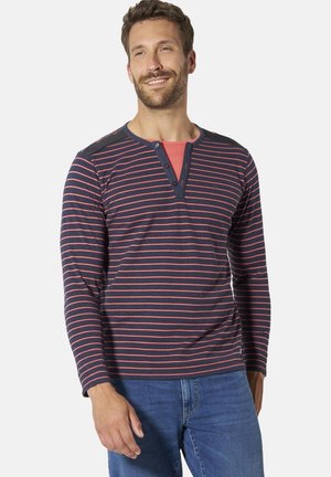 Smiling man wearing navy blue long-sleeve shirt with red horizontal stripes and jeans, standing against a plain light background.