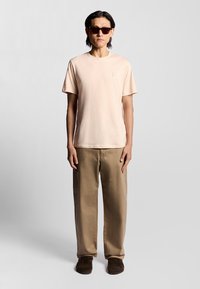 Lichtroze katoenen t-shirt, korte mouwen, ronde hals, geborduurd logo. Beige broek, relaxte pasvorm, effen structuur. Bruine instappers.