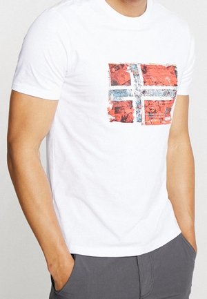 Mies, jolla on valkoinen t-paita, jonka rinnassa on kuluneen näköinen Norjan lipun kuvio, kädet harmaiden housujen taskuissa.