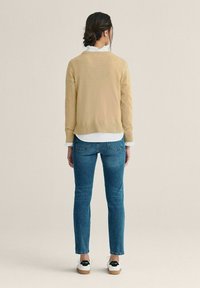 Beige neulepaita valkoisen paidan päällä, yhdistetty sinisiin skinny-farkkuihin ja valkoisiin kenkiin mustilla yksityiskohdilla, nähtynä takaa.