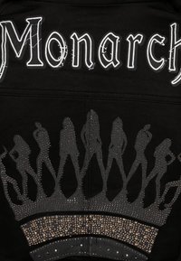Giacca nera con testo bianco "Monarch" e un design a silhouette sul fondo, arricchita da strass, creando un effetto testurizzato simile a una corona.