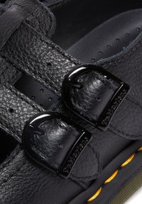 Chaussure en cuir noir avec deux boucles réglables, coutures apparentes et détail de couture jaune sur la semelle.
