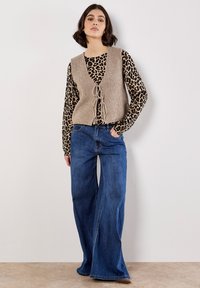 Beige knitted vest with open front and tie detail over a leopard print long-sleeve shirt, paired with wide-leg blue denim jeans.