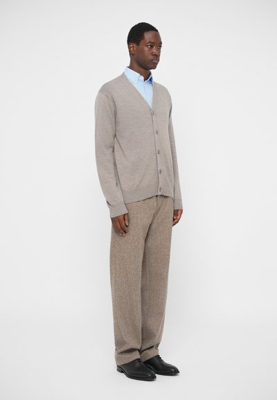 Άνδρας που στέκεται, φορώντας μπεζ κάρντιγαν, ανοιχτό μπλε πουκάμισο, χαλαρούς μπεζ παντελόνες και μαύρα ε�πίσημα παπούτσια σε λευκό φόντο.