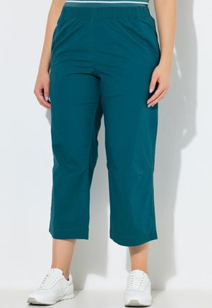 Türkisfarbene kurze Hose mit elastischem Bund und Seitentaschen, getragen mit weißen Turnschuhen an einer stehenden Person vor weißem Hintergrund.