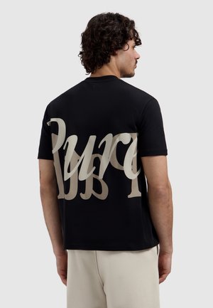 Man with curly dark hair wearing a black T-shirt with large beige script text on the back and light-colored pants, facing sideways.