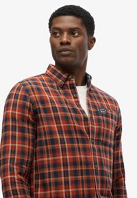 Red and blue plaid shirt with a button-down collar, featuring a textured fabric and a small logo patch on the chest.