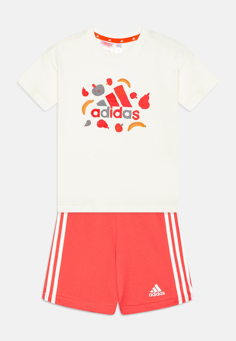 T-shirt bianco in cotone con design di frutta multicolori e logo "adidas"; abbinato a pantaloni corti rossi in cotone con strisce bianche e logo.