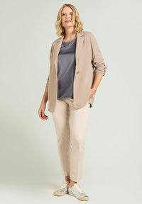 Beige Blazer über einem grauen Oberteil, kombiniert mit hellbeigen Hosen und silbernen Sneakers. Glatter Stoff, taillierte Passform und minimalistisches Design.