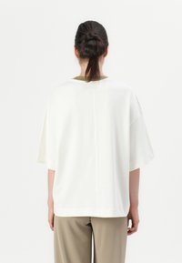 Persoon met donker haar in een knot, gekleed in een losse witte top met korte mouwen en beige broek, staand tegen een effen witte achtergrond.