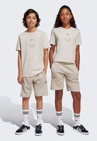 Μπεζ βαμβακερές μπλούζες και σορτς με διακριτικό λογότυπο στο κέντρο. Κάλτσες ριγέ σε μαύρο και άσπρο με μαύρα αθλητικά παπούτσια. Απλό φόντο.