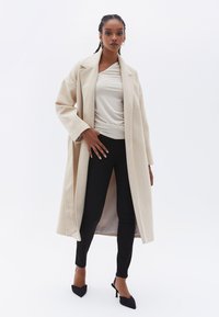 Abrigo de lana beige con solapas anchas, frente abierto y mangas largas, combinado con una blusa ligera y pantalones negros ajustados, además de zapatos de tacón puntiagudos negros.