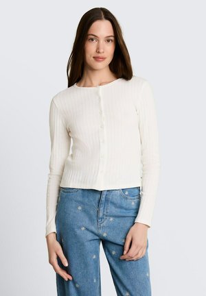 Vrouw met lang bruin haar draagt een witte geribbelde trui met knopen en een blauwe spijkerbroek met kleine witte geborduurde bloemmotieven.