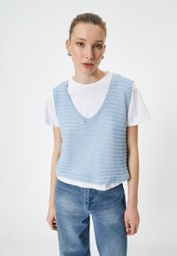 Chaleco de punto azul claro con escote en V, que presenta un patrón texturizado. Llevado sobre una camiseta blanca y combinado con unos jeans de mezclilla azul claro.