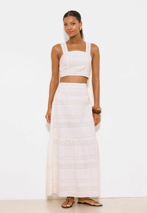 Vrouw draagt een wit mouwloos cropped topje en een bijpassende lange rok met oogjes, sandalen en houten armbanden, staand tegen een witte muur.