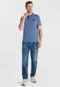 Polo shirt azzurro chiaro con un logo nero, abbinato a jeans blu sbiaditi con grandi tasche. Le scarpe sono sneaker grigio chiaro.