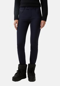 Tmavomodré úzké kalhoty s hladkou strukturou, obsahující kapsu na zip a zapínání na knoflík, doplněné černými zimními botami.