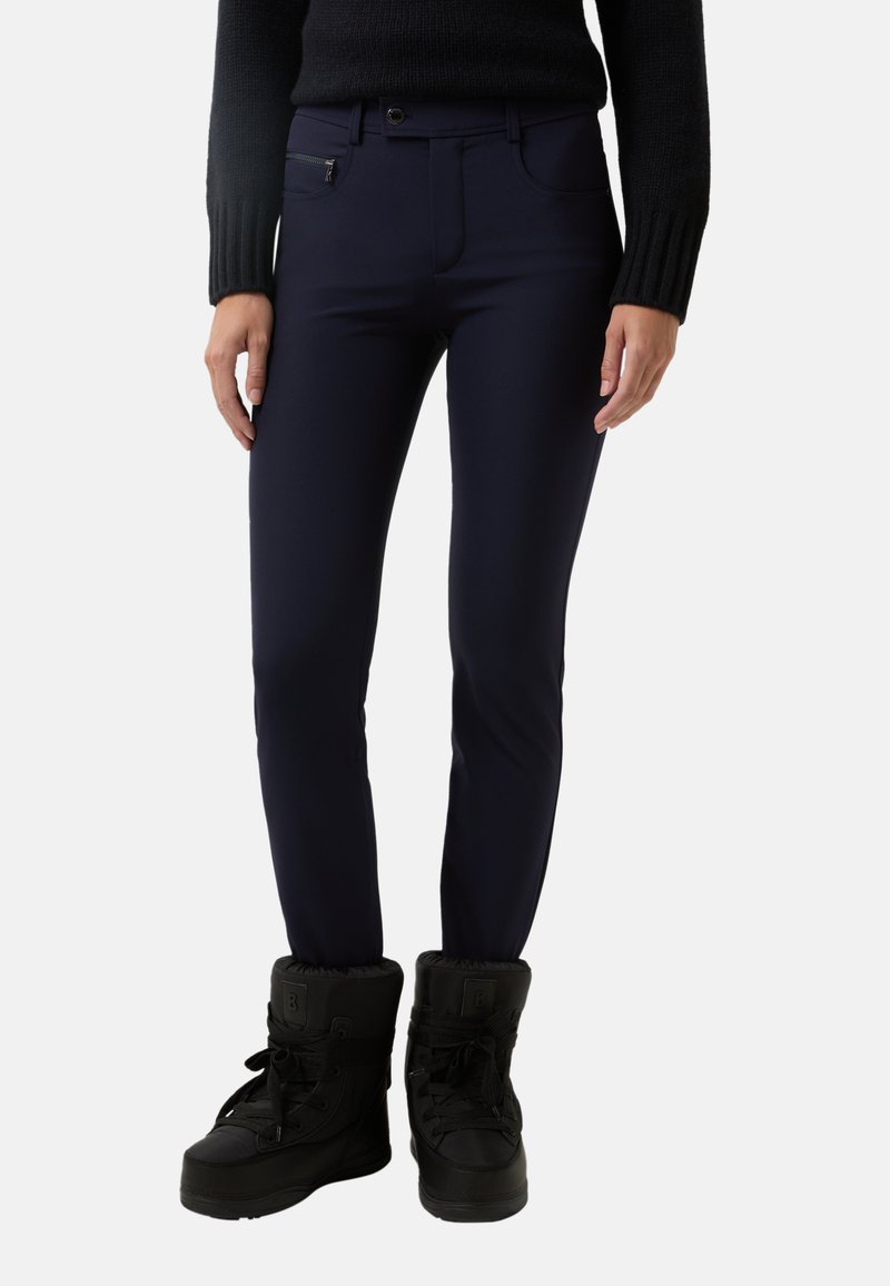 Tmavomodré úzké kalhoty s hladkou strukturou, obsahující kapsu na zip a zapínání na knoflík, doplněné černými zimními botami.