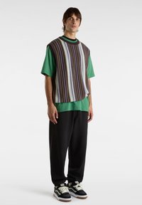 Gilet en tricot à rayures marron, violet et bleu superposé sur un t-shirt vert. Un pantalon de jogger noir et des chaussures noires complètent la tenue.
