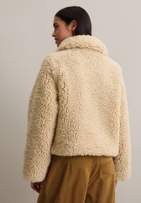 Veste en fausse fourrure beige avec une surface douce et texturée, dotée d'un large col et d'une coupe structurée, assortie à un pantalon en velours côtelé marron.
