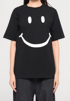 Persona que lleva una camiseta negra de gran tamaño con una gran cara sonriente blanca en el frente, combinada con vaqueros oscuros.