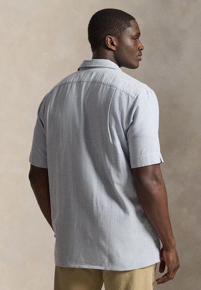 Moški, obrnjen stran, oblečen v svetlo modro srajco s kratkimi rokavi in tistmi tankimi navpičnimi črtami ter bež hlače, stoji pred nevtralnim ozadjem.