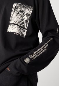 Zwarte longsleeve shirt met een textuur in het witte grafische ontwerp en geprinte adresdetails op de mouw, met een QR-code nabij de manchetten.