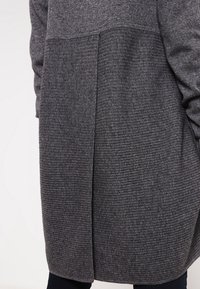 Cardigan en maille grise présentant un mélange de textures lisses et côtelées, avec un ourlet arrondi et une fente au dos pour plus de style et de confort.