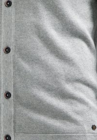 Grijze gebreide cardigan met zwarte knopen, een gladde textuur en een subtiele logoaccent bij de zoom. Gericht op praktische bruikbaarheid en comfort.