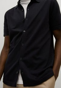 Musta lyhythihainen nappipaita, jossa on kudottu pinta, kaulus ja useita nappeja edessä. Päällä valkoinen paita.