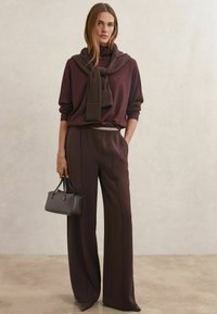 Pulover maro tricotat cu o eșarfă cu dungi purtată pe umeri, asortat cu pantaloni maro cu talie înaltă și croială largă și o geantă mică.