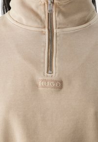 Sweat-shirt beige à fermeture éclair avec col montant, fermeture éclair argentée visible, et petit patch rectangulaire avec le mot "HUGO" brodé sous la fermeture éclair.
