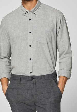 Mand iført lysegrå skjorte med knapper og opsmøgede ærmer samt mørkegrå ternede bukser, hænderne i lommerne.