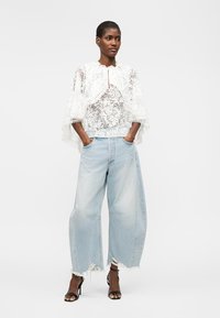 Žena v bielom čipkovom cape tope, širokých svetlomodrých rifliach s roztrhaným vzhľadom a čiernych sandáloch na vysokom opätku, stojaca s rukami v vreckách.