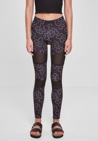 Crne legging hlače s bijelim cvjetnim uzorkom, s mrežastim panelima na gornjim nogama i visokim strukom. Model nosi crne sandale.