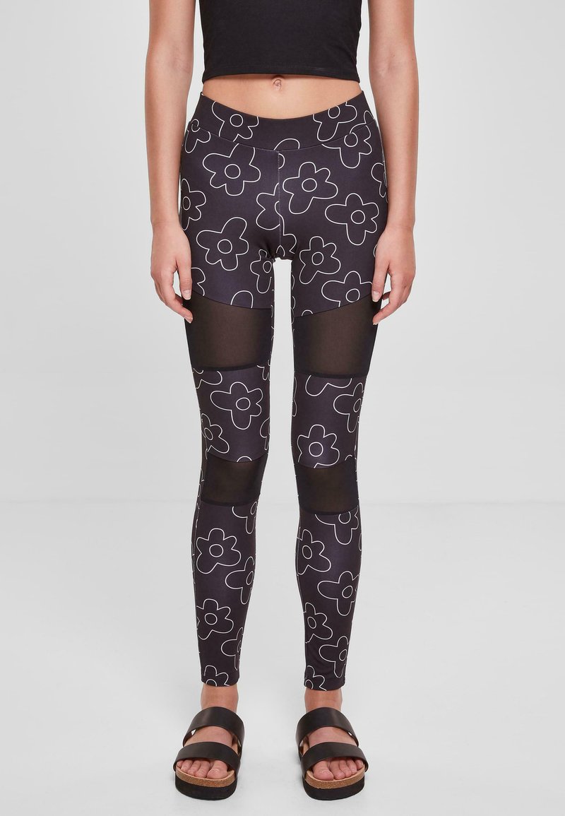 Crne legging hlače s bijelim cvjetnim uzorkom, s mrežastim panelima na gornjim nogama i visokim strukom. Model nosi crne sandale.