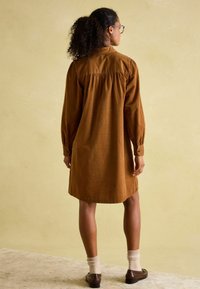 Braunes Cordhemdkleid mit langen Ärmeln, gerafftem Rückendetail und Knopfmanschetten, kombiniert mit hellen Stricksocken und dunklen Schuhen.